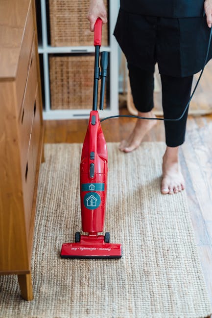 A person using a yellow and black vacuum cleaner on a patterned carpet in a well-lit room. The individual is kneeling and inserting a cloth into the vacuum's dust container, demonstrating surface clea